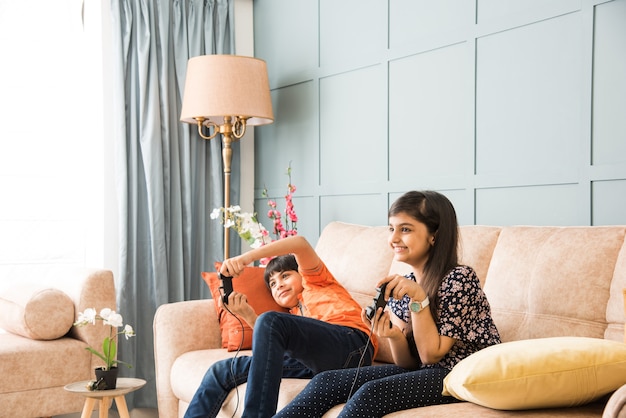 Cheerful Asian Indian kids playing video game using controller or joystick, sitting on sofa, couch