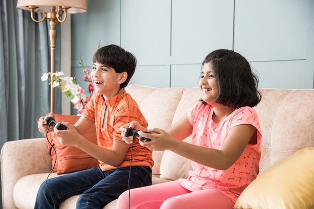 Cheerful Asian Indian kids playing video game using controller or joystick, sitting on sofa, couch