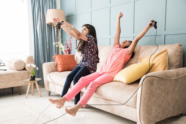 Cheerful asian indian kids playing video game using controller\
or joystick, sitting on sofa, couch