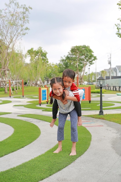 陽気なアジアの小さな男の子と女の子の子供が庭で背中に乗って遊ぶ兄弟が妹の背中に乗っている