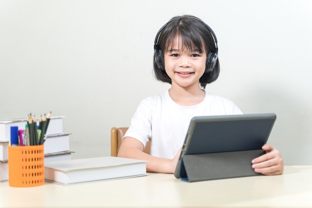 Cheerful Asian little girl student wears headphone writes on digital tablet to study at home