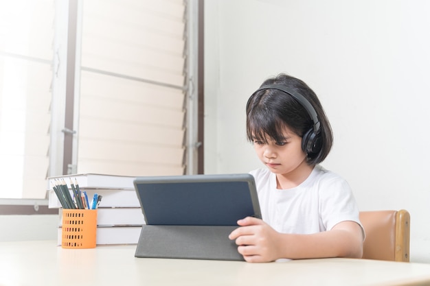 Cheerful Asian little girl student wears headphone writes on digital tablet to study at home