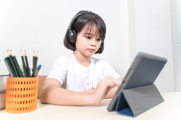 Cheerful Asian little girl student wears headphone writes on digital tablet to study at home