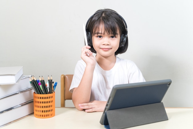 Cheerful Asian little girl student wears headphone writes on digital tablet to study at home