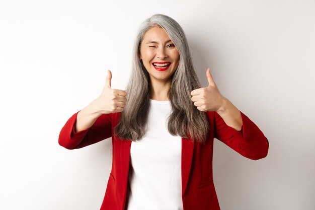 Cheerful asian mature woman winking, smiling pleased and showing thumbs-up in approval, like and agree, standing against white background