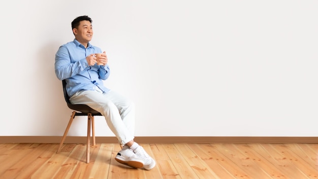 Cheerful asian middle aged man sitting on chair at home drinking herbal tea or coffee looking at