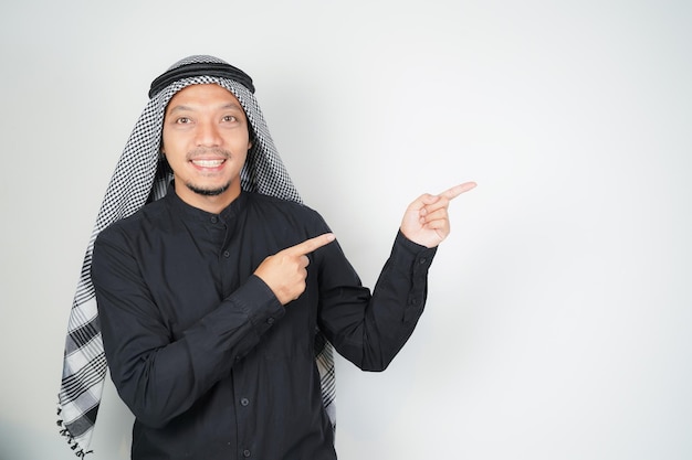 Cheerful Asian Muslim man wearing Arab turban sorban pointing hand finger at empty space on isolated