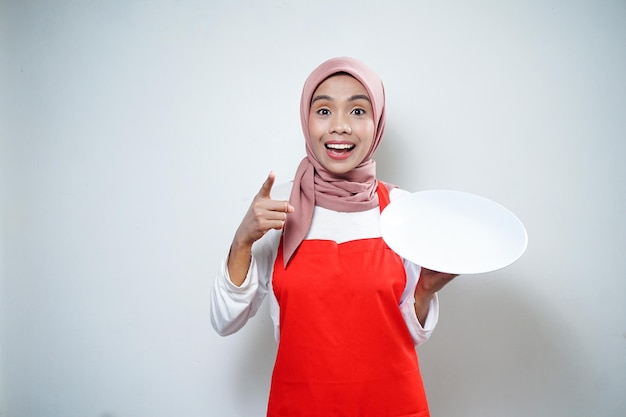 空のプレートを保持している赤いエプロンで陽気なアジアのイスラム教徒の女性食品広告料理のコンセプト