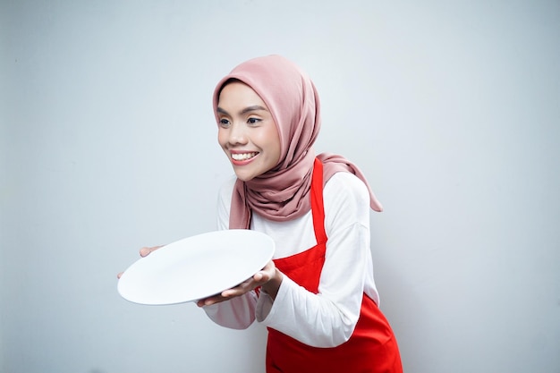 空のプレートを保持している赤いエプロンで陽気なアジアのイスラム教徒の女性食品広告料理のコンセプト
