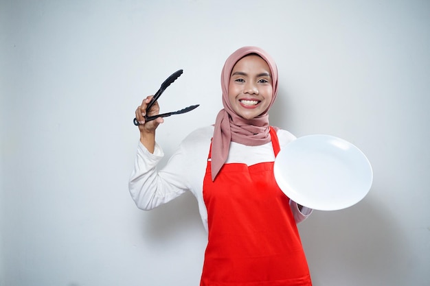 Cheerful asian muslim woman in red apron holding empty plate and tongs