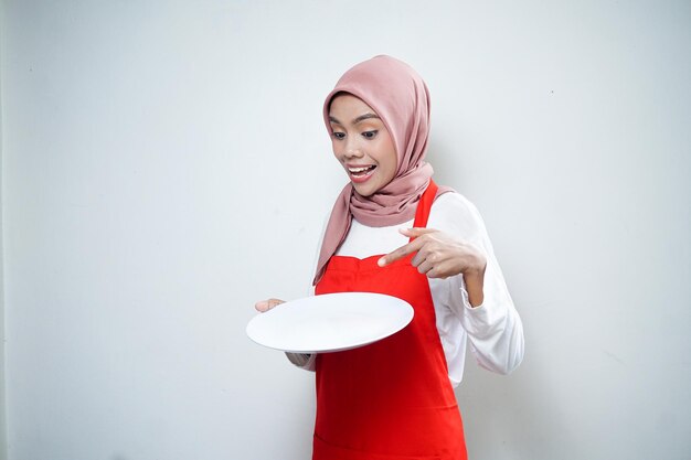 cheerful asian muslim woman in red apron pointing at empty plate food advertisement cooking concept