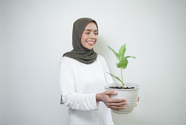 Cheerful Asian muslim woman in tshirt and hijab holding plant isolated on white background