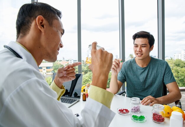 Cheerful Asian nutritionist in medical uniform smiling and demonstrating vitamin pills to man while sitting at table during work in hospital