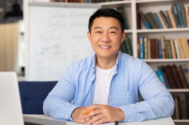 Photo cheerful asian teacher man smiling sitting near laptop at workplace