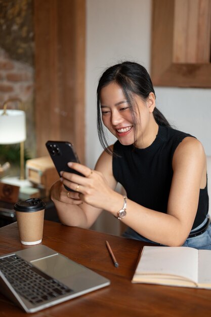 陽気なアジア人女性が、カフェに座りながら携帯電話でオンラインの面白い動画を楽しんでいます