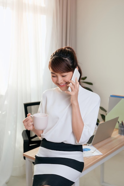 携帯電話で会話をし、オフィスでコーヒーを飲む陽気なアジア人女性