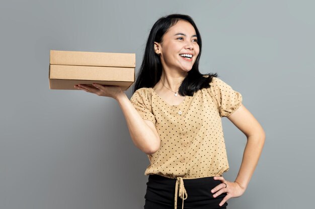 Cheerful asian woman holding cardboard box with one hand on waist