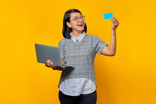 Cheerful asian woman holding laptop and looking at credit card over yellow background