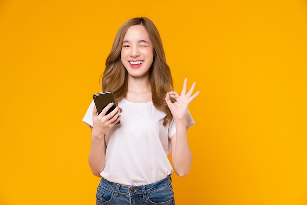 Cheerful asian woman holding smartphone and shows ok sign on light yellow background.