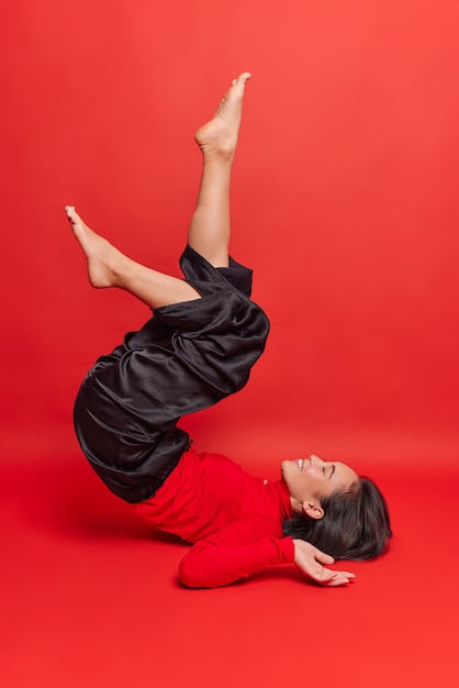 Жизнерадостная азиатская женщина держит ноги поднятыми, пытается встать на плечи, позирует босиком, носит водолазку и черные брюки, делает упражнения после работы, изолирована от красной стены, будучи в хорошей форме