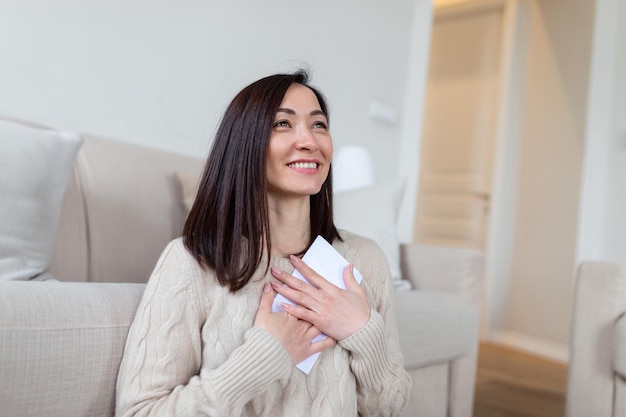 Cheerful asian woman presses received letter to the chest at\
homer receiving invitation or good news about approved loan\
mortgage tax insurance getting notice about acceptance of\
statement