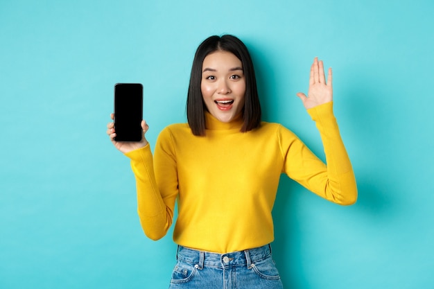 Donna asiatica allegra che mostra lo schermo vuoto dello smartphone e alza la mano per salutarti, salutarti, in piedi su sfondo blu.