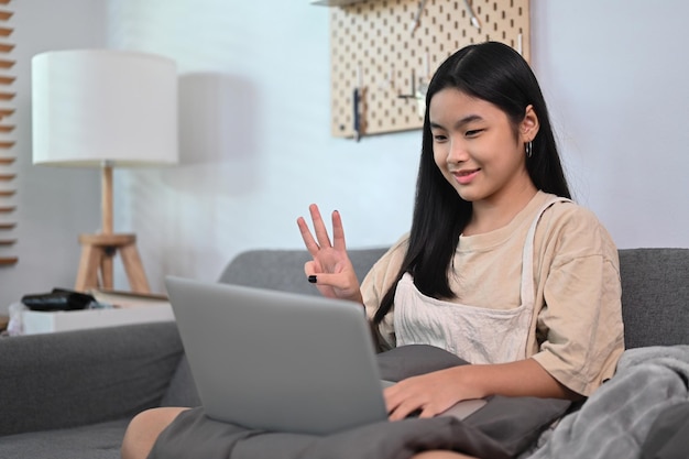 Donna asiatica allegra che si siede sul divano e che ha una videochiamata con un computer portatile