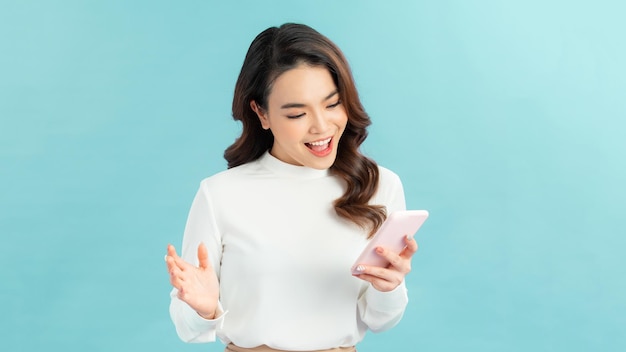 Cheerful asian woman standing isolated over blue background holding mobile phone celebrating