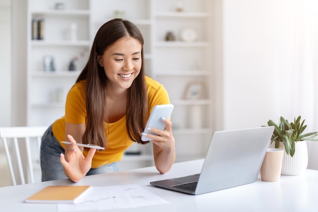 Cheerful asian woman using smartphone while working at desk in home office