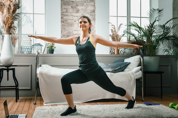 A cheerful athlete with black  hair lunges in the bedroom with online training