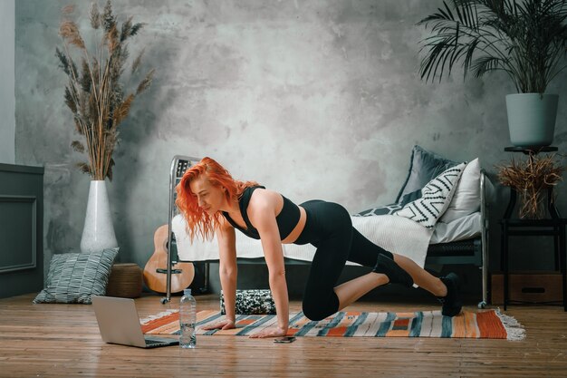 A cheerful athlete with blond  hair lunges in the bedroom with online training.