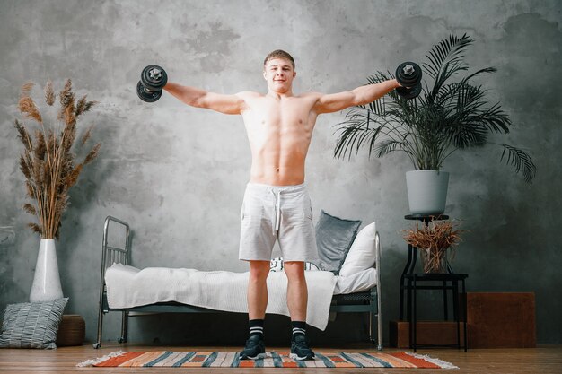 A cheerful athlete with blond hair swings to the side with dumbbells  in the bedroom. The young man goes in for sports at home.