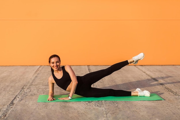 Cheerful athletic girl in tight sportswear, black pants and top, practicing yoga, doing side plank pose with leg lift, stretching muscles, training flexibility. health care, sport activity outdoor