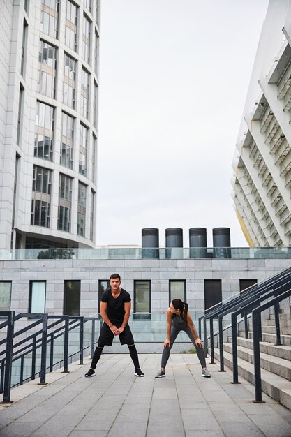 Cheerful athletic man and woman are bending for stretching while training on stairs among urban building