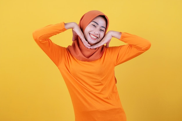 Cheerful attractive asian woman demonstrates copy space on blank yellow wall, has happy friendly expression, dressed casually wearing hijab, poses indoor