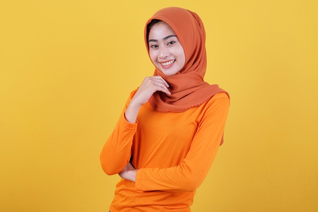 Cheerful attractive asian woman demonstrates copy space on blank yellow wall, has happy friendly expression, dressed casually wearing hijab, poses indoor