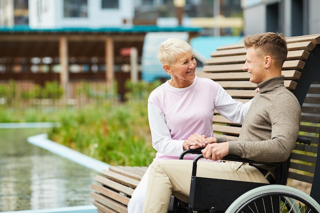 Cheerful attractive blond woman sitting on wooden bench outdoors and chatting with young handicapped