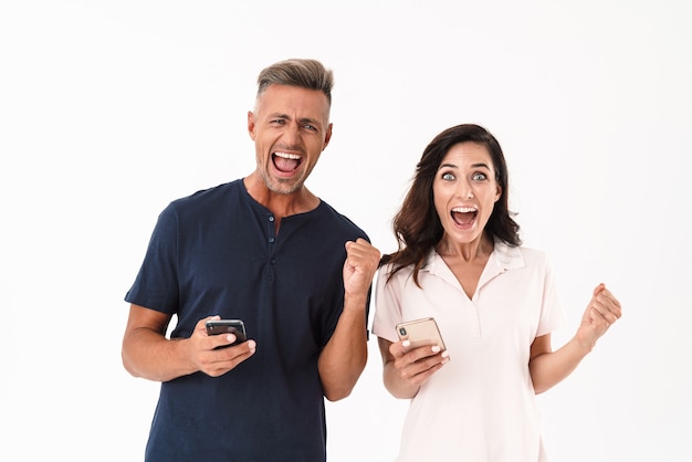 Cheerful attractive couple wearing casual outfit standing isolated over white wall, using mobile phone, celebrating success