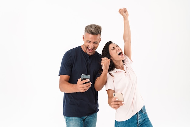 캐주얼 복장을 하고 흰 벽에 격리된 채 휴대폰을 사용하여 성공을 축하하는 쾌활한 매력적인 커플