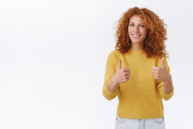 Cheerful attractive curly ginger woman in yellow sweater show thumbsup in approval cheering or supporting you as give positive feedback recommend product agree like event white background