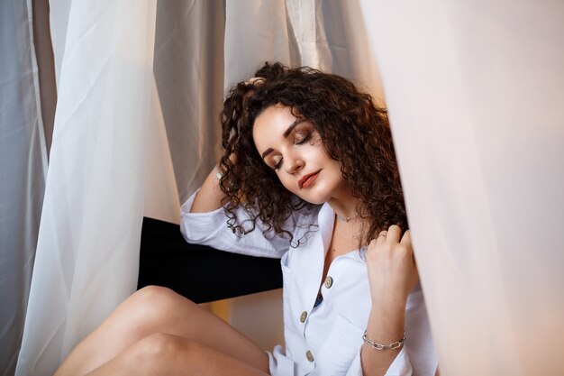 A cheerful, attractive girl sits on a chair, an armchair with a smile in a white shirt, sexy, cute, holds her hair