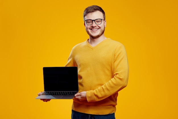 Foto uomo allegro e attraente in maglione giallo che mostra un nuovo moderno dispositivo portatile sullo sfondo arancione