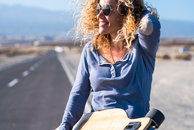 Allegra attraente bella donna adulta sorridere e godersi la libertà utilizzando il lungo tavolo da skateboard con la strada sullo sfondo - concetto di viaggio e godersi l'attività leiusre