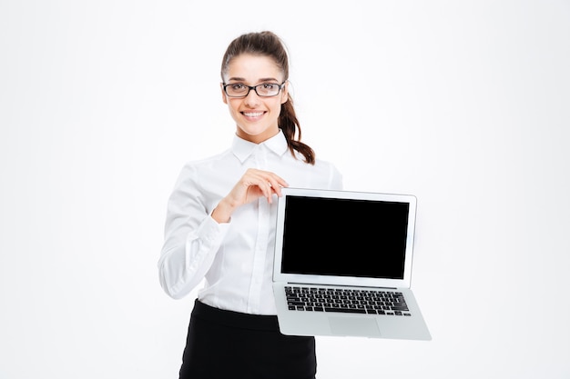 Cheerful attractive young businesswoman in glasses holding blank screen laptop over white wall