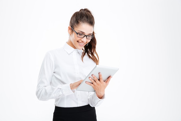 Cheerful attractive young businesswoman in glasses standing and using tablet over white wall