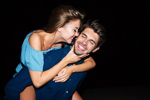 Cheerful attractive young man holding girlfriend on his back at night outdoors