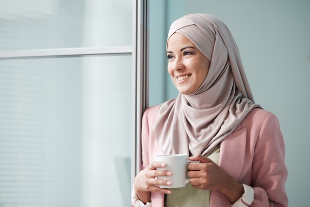 문에 서서 멀리 보면서 찻잔을 들고 분홍색 hijab에 쾌활한 매력적인 젊은 무슬림 여성