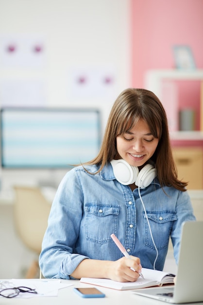 Cheerful attractive young woman in denim shirt wearing wired headphones on neck sitting at desk and