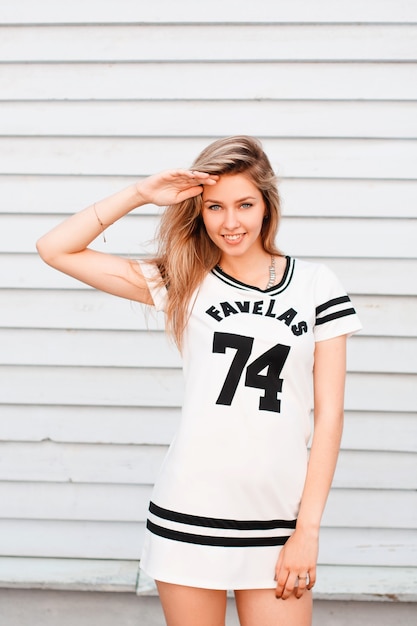 Cheerful attractive young woman with a beautiful smile in a sporty white summer dress with a black pattern near a wooden vintage building on a warm summer day. Happy girl enjoys the weekend.