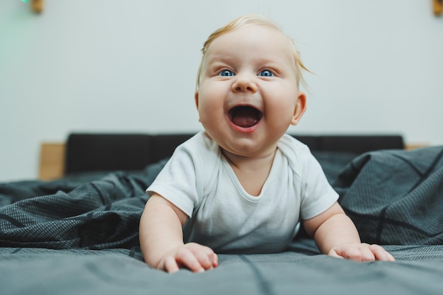 A cheerful baby boy with beautiful big eyes is lying on the bed at home in a white bodysuit on gray bedding The child is lying in bed at home